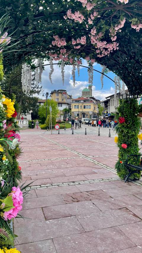 A lively town square framed by flower garlands bustles with people under a distant green-domed church.