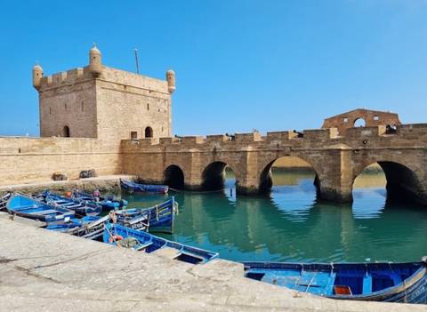 Stone battlements overlook a small harbour where vivid blue fishing boats float on turquoise water.