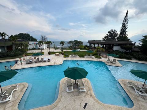 A resort pool overlooks misty Lake Atitlán and distant volcano peaks under a cloudy sky.