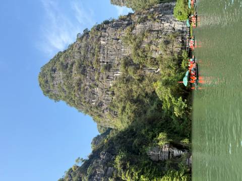 Tall green karst peak covered in vegetation reflecting in calm river water on a bright day.
