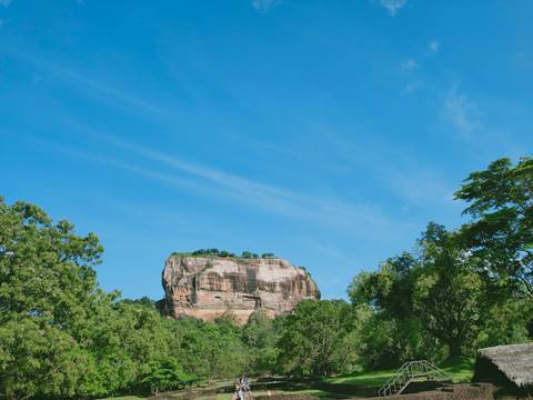 The towering granite monolith of Sigiriya rises above dense green forest against a vivid blue sky.