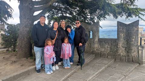 Smiling family group poses on a stone terrace overlooking distant rolling hills under a cloudy sky.