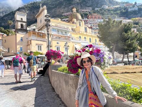 Traveler smiling beside vibrant flower display on pedestrian street in Positano with church dome behind.