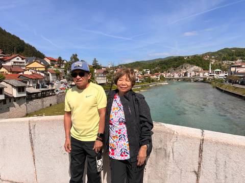 Couple on bridge with river and red-roofed town and forested hills behind.