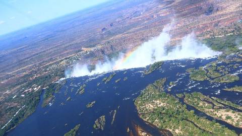 Oblique aerial of Victoria Falls with rising mist and a vivid rainbow cutting across the canyon.