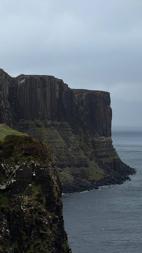 Moody sea cliff with sheer basalt walls plunging into grey North Atlantic waters under overcast sky.