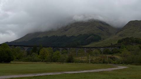 The Glenfinnan Viaduct arcs across a green valley beneath mist-shrouded highland mountains and low gray clouds.