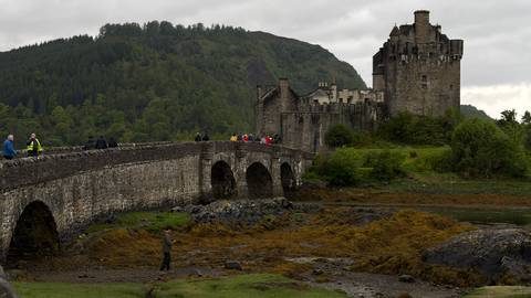 Eilean Donan Castle stands on a small island linked by an old stone bridge, framed by rugged hills and tidal shore at low tide.