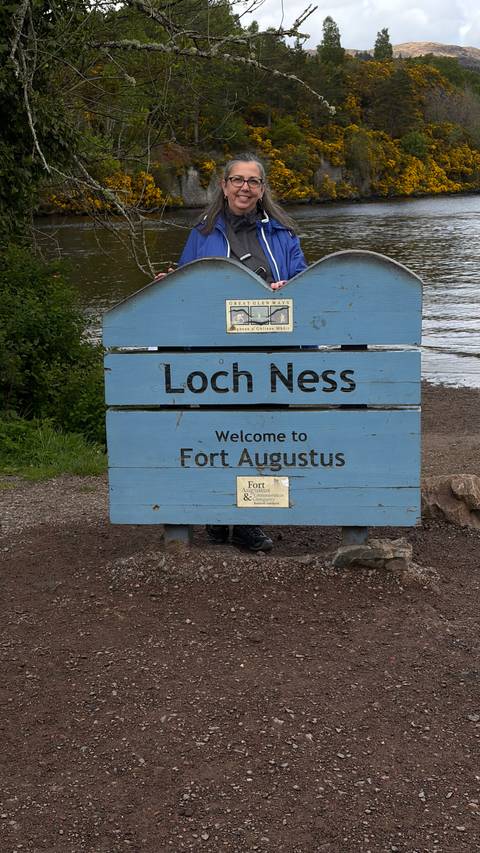 A blue wooden sign reads "Loch Ness – Welcome to Fort Augustus" by the lakeshore.