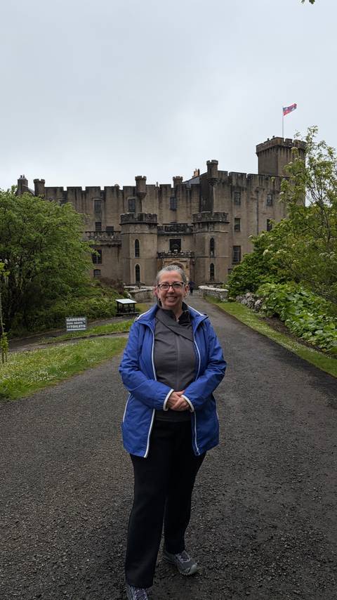 A smiling traveller in a blue rain jacket poses on a driveway leading to a historic Scottish castle surrounded by lush gardens.