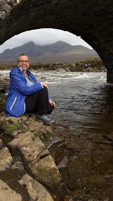 Woman in blue rain jacket sitting on riverside rocks beside fast-moving water