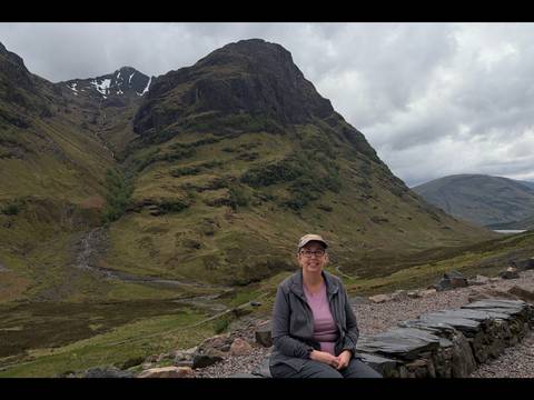Traveller seated on stone wall with dramatic green mountains of Glencoe rising behind under cloudy skies