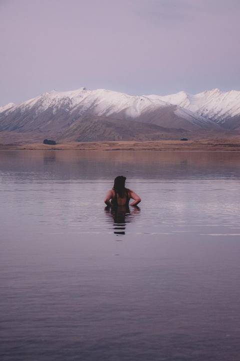 Woman standing waist-deep in tranquil lake at dusk with mountains reflecting on calm water