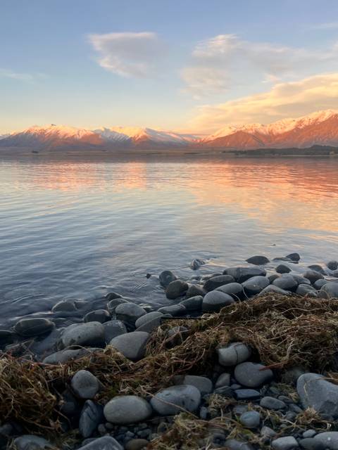 Soft golden reflections of sunset over lake with snow-tipped mountains and smooth stones in foreground