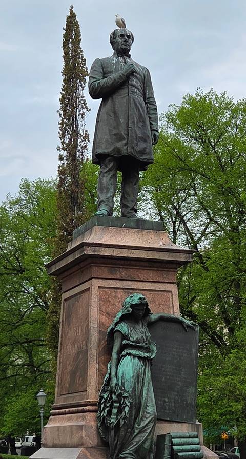 Base of a bronze statue atop a granite pedestal surrounded by spring foliage in a city park