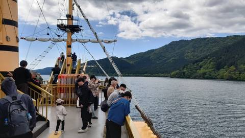 Tourists standing on the deck of a yellow sightseeing boat cruising a serene lake surrounded by forested mountains.