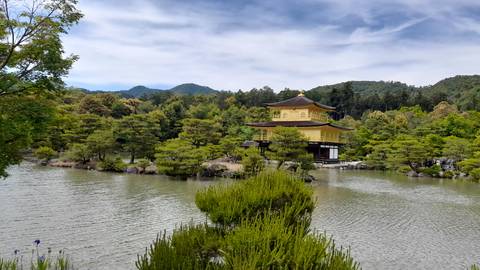 Golden Pavilion (Kinkaku-ji) reflecting over a tranquil pond surrounded by manicured pines under blue sky.