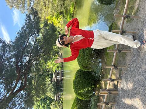 Visitor waves cheerfully in a lakeside Japanese garden surrounded by lush greenery.