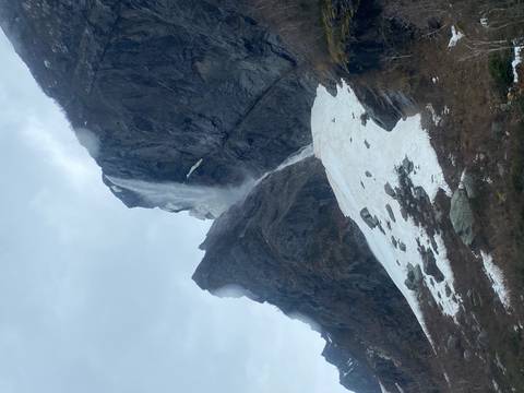 Tall waterfall plunges down a sheer cliff beside lingering spring snow and misty skies.