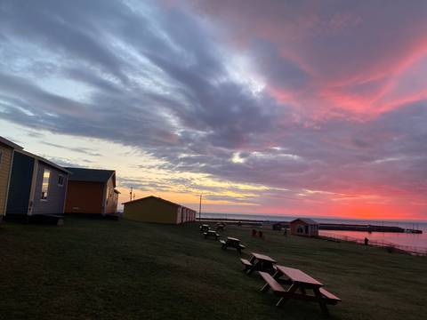 Vibrant sunset colours paint the sky above coastal cabins and a small breakwater.