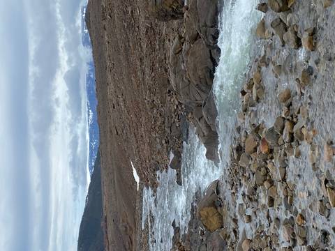 Fast-flowing rocky river cuts through a barren tundra with distant snowy mountains under grey skies.