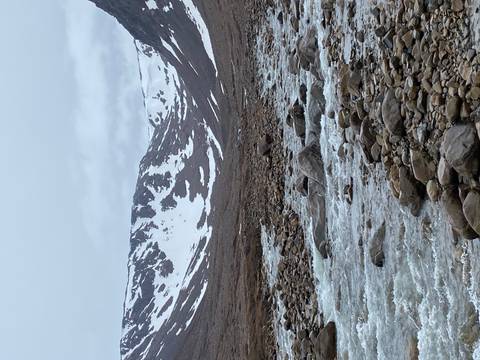 Snow-dusted valley strewn with boulders and a rushing melt-water stream under grey skies.