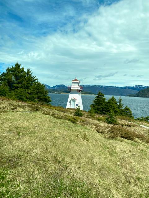 A small white and red lighthouse stands on a grassy hill overlooking a calm lake and distant snow-capped mountains beneath a partly cloudy sky.