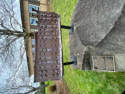 A riveted metal beam displayed on supports with a tree and building in the background; a small plaque rests on a large stone in front.