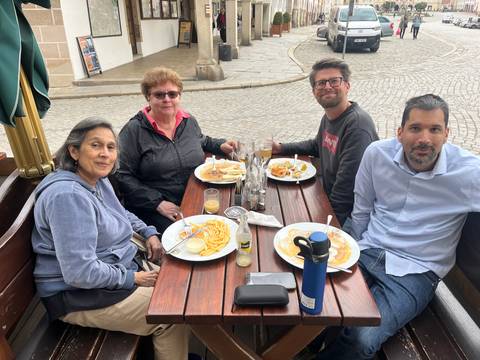 Four friends enjoy plates of Czech food and beers at an outdoor wooden table on a cobblestone street.