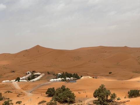 White desert camp tents cluster at the base of vast orange sand dunes under a hazy sky.