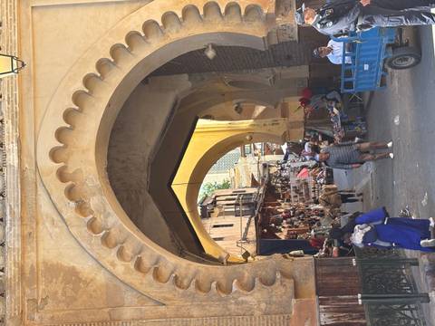 A bustling archway in the Fes medina filled with shoppers, stalls and carts beneath aged yellow stonework.