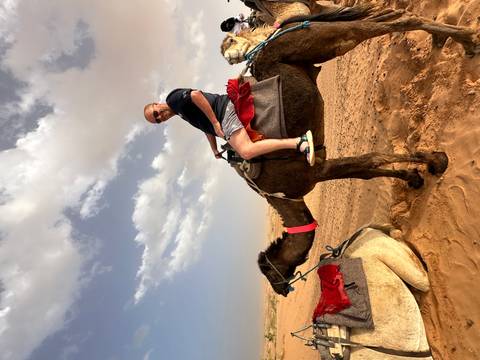 A smiling traveler rides a camel across reddish desert sands beneath scattered clouds.
