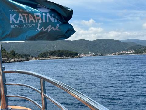View from a boat railing toward forested Adriatic islands and a distant coastal village with a Katarina Line flag in the foreground.