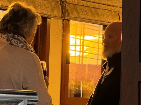 Two older travelers chat over glasses of wine in a cabin as warm sunset light pours through the window.