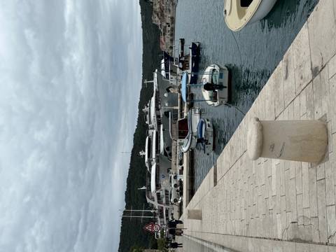 Small marina with moored yachts and tenders under a cloudy sky, stone quay in the foreground.