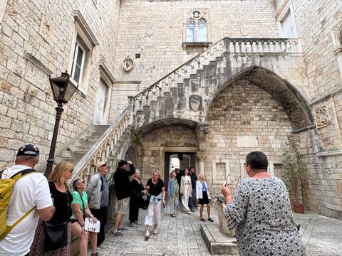 Guided tourist group stands in a medieval stone courtyard beneath an ornate arched staircase.