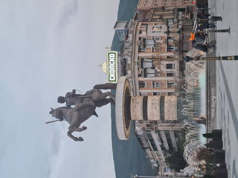 Equestrian statue fountains and neon signage dominate a busy city square at dusk in Skopje.