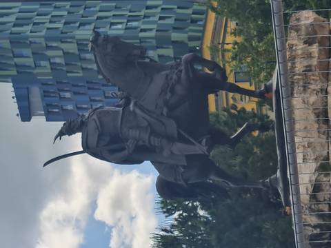 Dark bronze equestrian statue of a warrior wielding a curved sword standing before modern buildings.