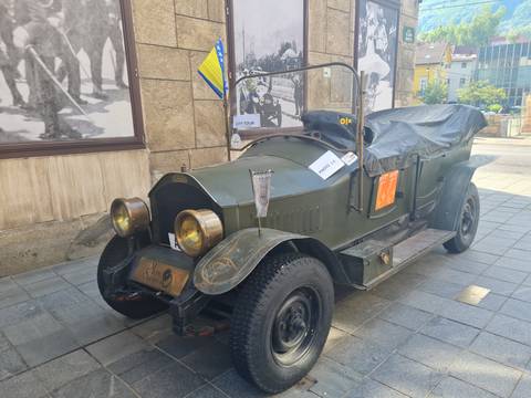 Early 20th-century touring car parked on a city sidewalk with informational placards on its hood.