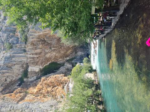 Clear turquoise river emerges from a cavern at the base of a towering limestone cliff beside riverside cafés and trees.