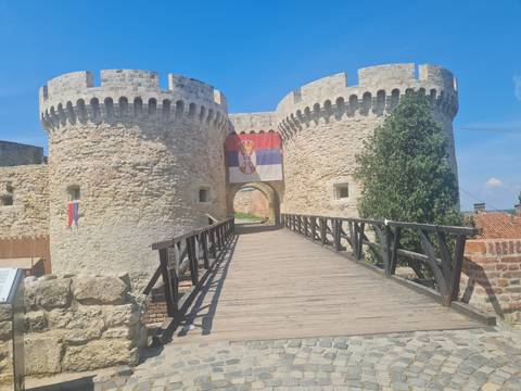 Stone bridge leading to round towers of a medieval fortress bearing the Serbian flag under blue sky.