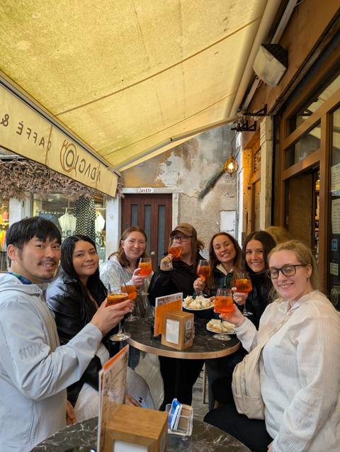 Seven young travelers raise Aperol spritz cocktails and smile inside a cozy Italian bar with rustic walls.