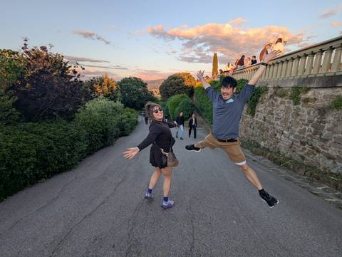 Two travellers joyfully jumping on a hillside lane with sunset clouds and a scenic city panorama behind them.