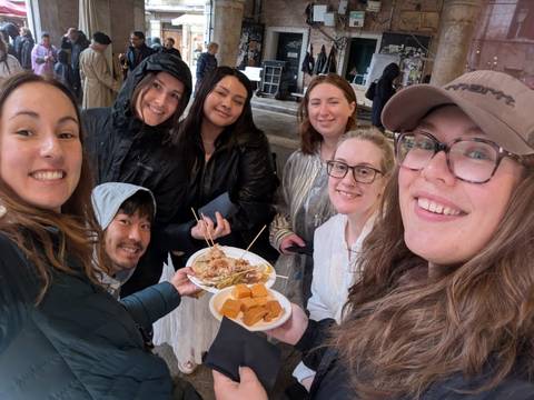 Six travellers gather under shelter, holding plates of street food and smiling toward the phone camera.