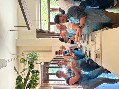 Cooking class scene with eight participants in blue aprons proudly holding freshly made pasta inside a bright studio.