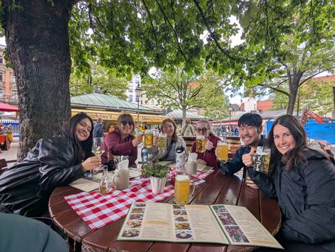 Friends raise large beer mugs and wine glasses at an outdoor Bavarian beer garden beneath leafy trees.