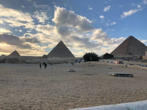 Wide desert scene with the three Pyramids of Giza rising behind a few visitors under a dramatic partly-cloudy sky.
