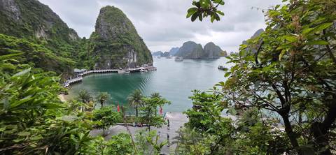 Lush green limestone islands and anchored boats in an emerald bay viewed through tropical foliage.