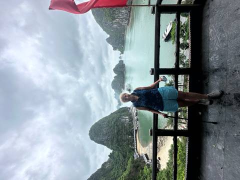 Older traveller with a cane poses on a balcony overlooking the limestone islands of Ha Long Bay.