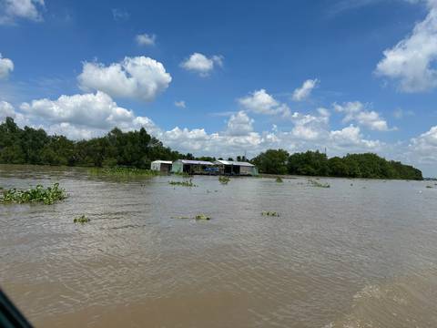 Wide muddy river scene with floating vegetation and a few stilted houses under a bright blue sky.